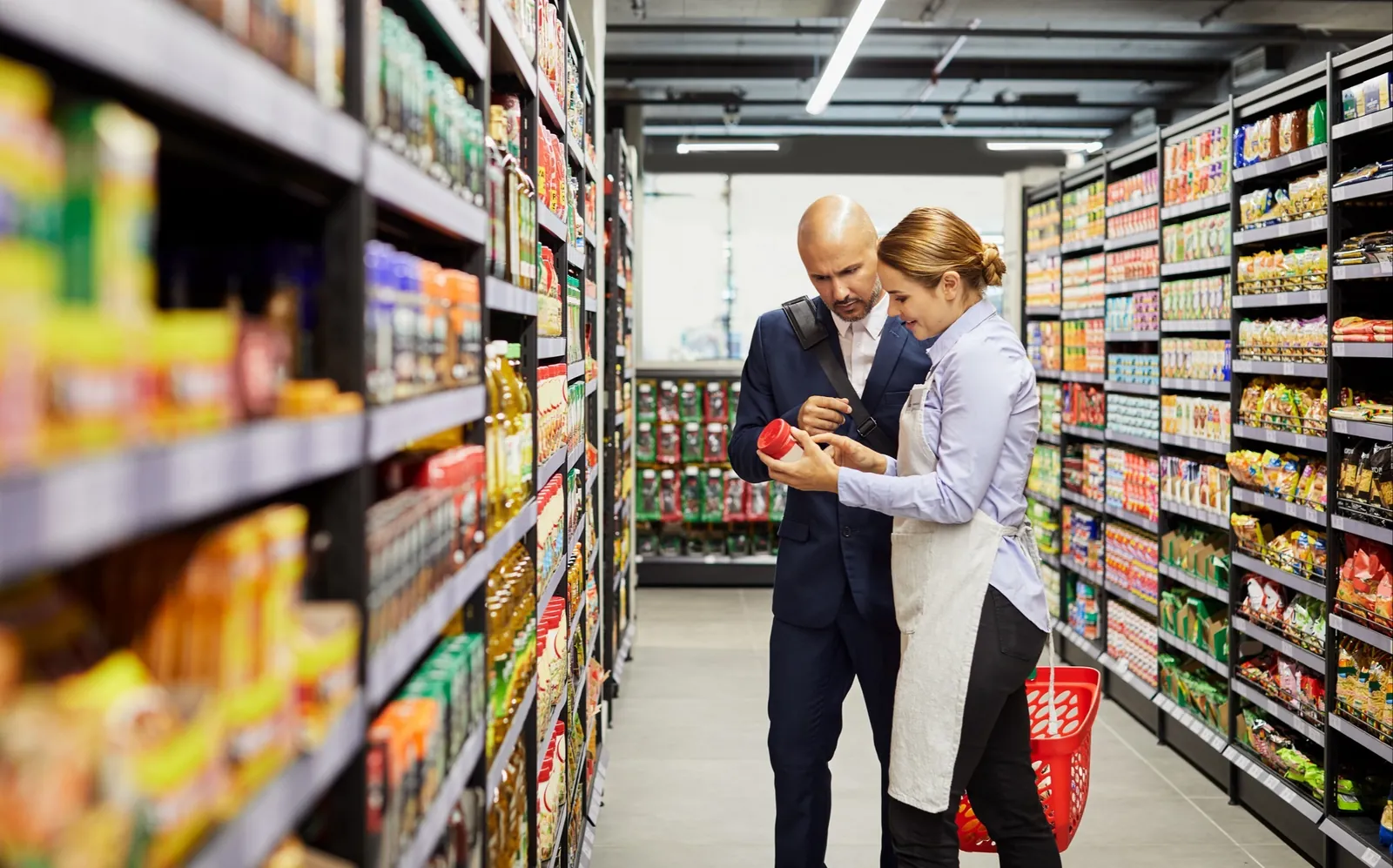 Shop assistant helping a customer read a product label in a convenience store aisle