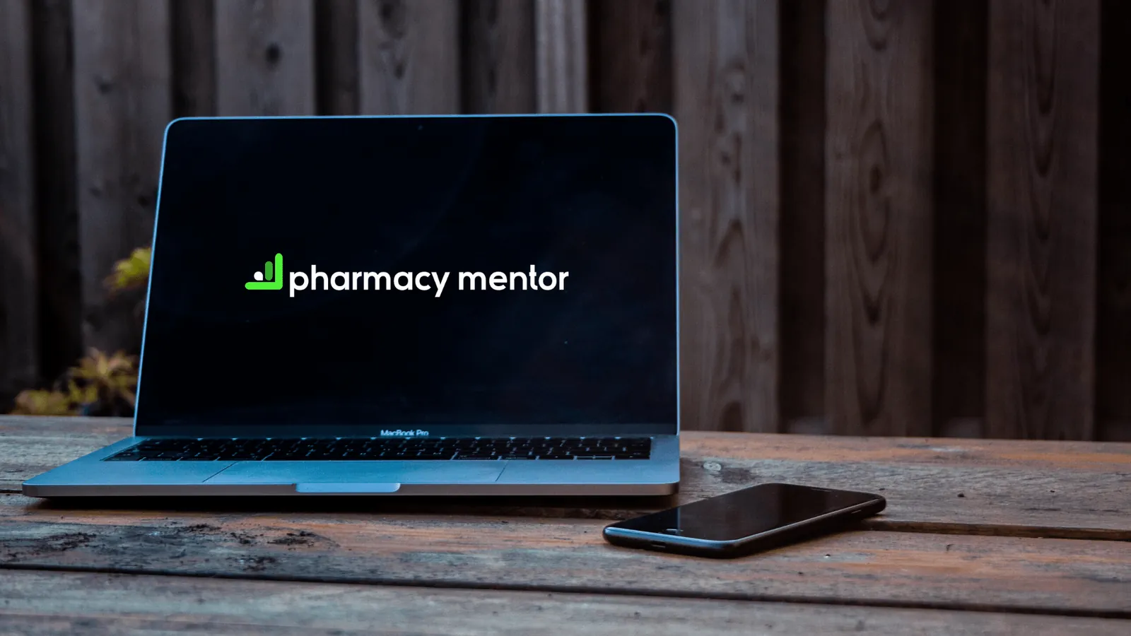 Laptop showing the Pharmacy Mentor logo on a wooden table next to a phone